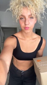 Must silmapaistva laia rihmaga must crop top, mille alla on sobitatud kõrge vöökohaga mustad püksid. Esiplaanil on pappkast, millel on kiri "Armastus". Taustal on lihtne siseruum.