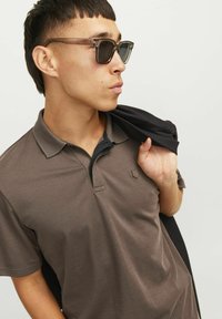 Hnedé polokošeľa s textúrovaným golierom, na hrudi s malým emblémom. Čierna bunda je prehodená cez rameno. Sklá slnečných okuliarov sú priehľadné.