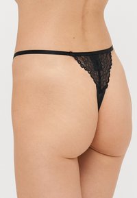 Sort blonde trusse med tyndt bånd, der har bølgede kanter og et gennemsigtigt design, som fremhæver intrikate blomstermønstre. Bagside vist.