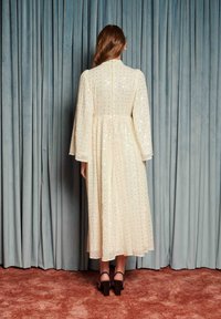 Žena v krémových dlhých rukávoch midi šatách s flitrovými detailmi stojí čelom k modrým závesom na hrdzavo sfarbenom koberci.