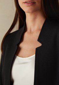 Czarna marynarka z otwartym kołnierzem, noszona na białym satynowym topie, podkreślająca gładkie tekstury i czyste linie.