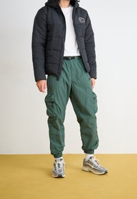 Black puffer jacket with hood and logo, white t-shirt, green cargo pants with drawstring, and gray sneakers. Textured materials and relaxed fit.