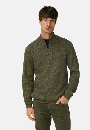 Mann in olivgrünem Strickpullover mit Viertelreißverschluss und dunkelgrüner Hose, steht mit einer Hand in der Tasche vor einem weißen Hintergrund.