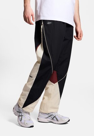 Mężczyzna ubrany w spodnie dresowe z blokami kolorów czarnego, beżowego i bordo oraz białe buty do biegania, stojący na jednolitym tle.