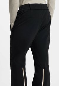 Czarne spodnie narciarskie z logo na pasku, posiadają boczne kieszenie oraz pionowe białe i złote paski przy mankietach. Gładka, teksturowana tkanina.