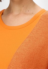 Orangenes Oberteil mit rundem Ausschnitt, auf der einen Seite mit Rippstruktur und auf der anderen Seite mit glatt gestricktem Muster, das einen visuellen Kontrast erzeugt.