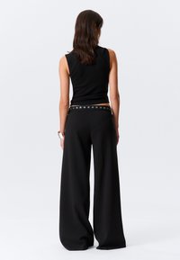 Haut noir sans manches associé à un pantalon noir à jambes larges, doté d'une taille ceinturée avec des détails circulaires en métal. Texture lisse et coupe ajustée.