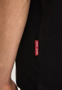 Czarny bawełniany t-shirt z widoczną czerwoną metką materiałową z napisem „JOOP! JEANS” na bocznym szwie. Gładka faktura, krótkie rękawy.