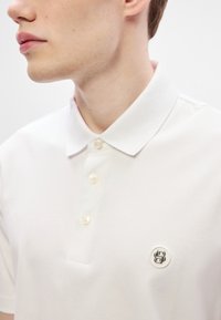 Λευκό πόλο από βαμβάκι με κλασικό γιακά, διαθέτει τρία κουμπιά και μικρό κεντημένο λογότυπο στο στήθος. Λιτή υφή.