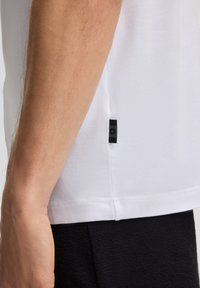 Witte T-shirt met korte mouwen en een gladde текстuur, voorzien van een zwart logo-label aan de zijkant, gecombineerd met zwarte textuurbroek.