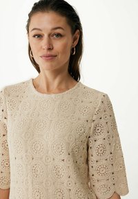 Beige spetsöverdel med rund halsringning och korta ärmar, med intrikata cirkulära mönster och en strukturerad yta.