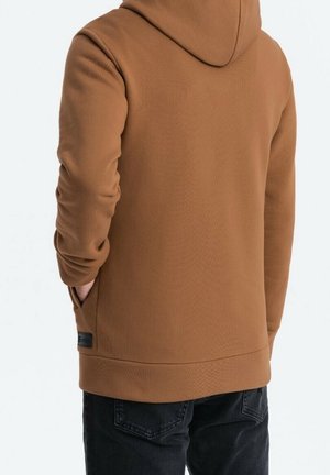Personne portant un sweat à capuche marron, les mains dans les poches, vue de dos, assorti à un jean foncé.