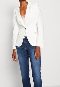 Blazer blanco con un diseño ajustado, cierre de un botón y detalles en dorado. Combinado con una blusa de encaje clara y jeans de mezclilla azules.