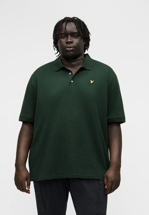 Μαύρος άνδρας με κοντά ντραντλοκ, φοράει σκουρό πράσινο πόλο μπλουζάκι με κοντά μανίκια και ένα μικρό κίτρινο λογότυπο στο αριστερό στήθος.