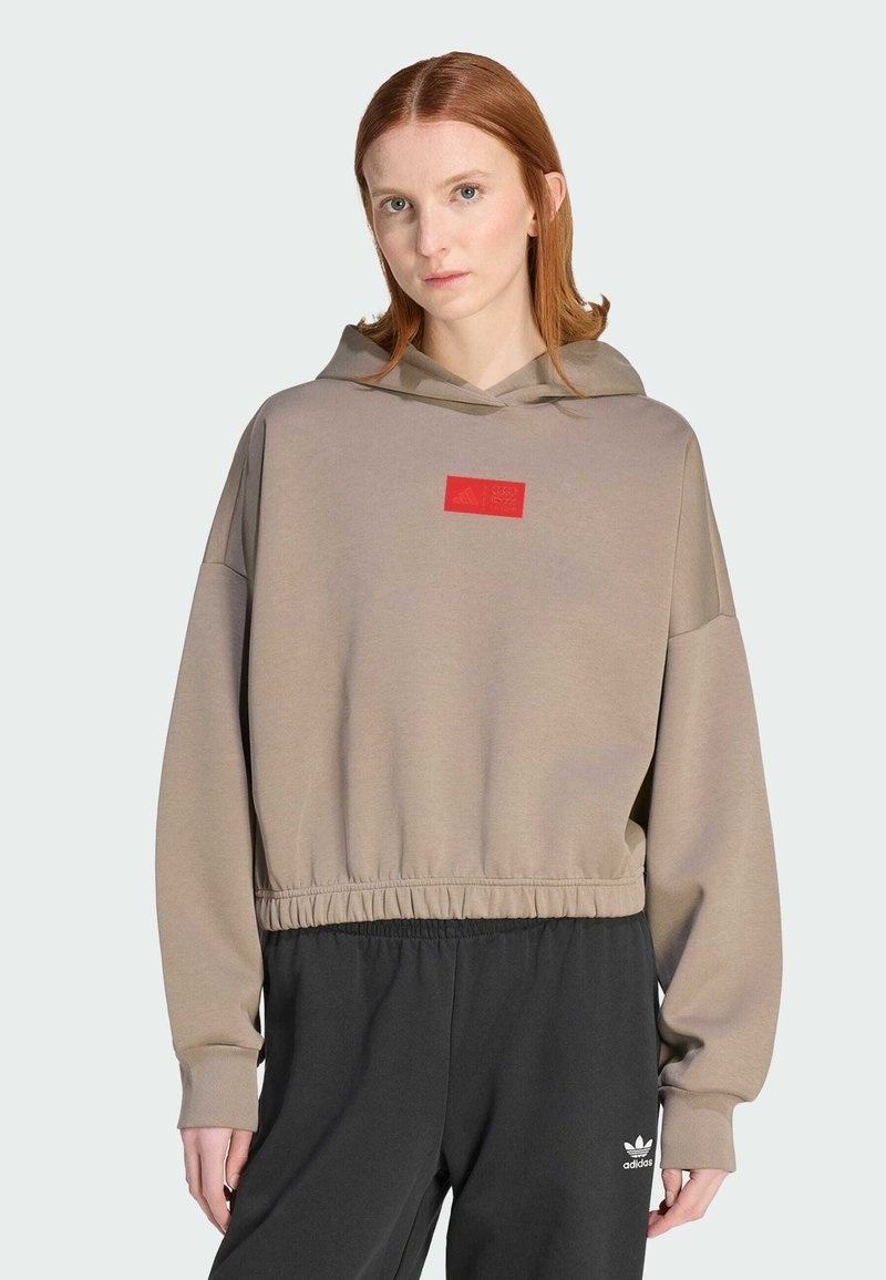 Woman with red hair wearing a beige cropped hoodie with a red logo and black Adidas pants with a white logo on a light background.