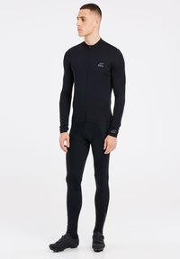 Maillot de cyclisme noir à manches longues avec un col haut et une fermeture éclair, associé à un legging noir ajusté et des chaussures de cyclisme noires.