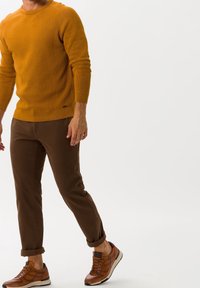 Mann in einem senfgelben Pullover, braunen hochgekrempelten Hosen und braunen Ledersneakern, der vor einem schlichten weißen Hintergrund steht.