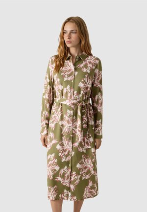 Žena oblečená v olivově zelených midi šatech-košili s růžovým a bílým květinovým potiskem a zavazovacím páskem, stojící před jednobarevným světle šedým pozadím.