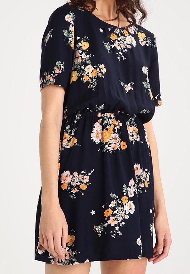Femme portant une robe bleu marine à manches courtes avec des motifs floraux orange, blanc et rose, et une taille élastique.