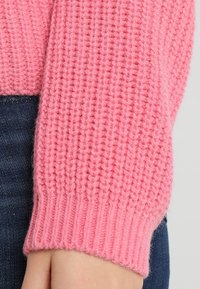 Gros plan sur une main portant une manche de pull en tricot rose sur un jean en denim bleu foncé.