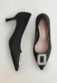 Chaussures à talons hauts en suède noir avec un bout pointu et une boucle décorative en rhinestones sur chaque zone des orteils. Doublure intérieure lisse ; talon fin.