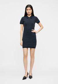 Γυναίκα στέκεται μπροστά σε λευκό φόντο φορώντας φόρεμα πλεκτό κοντό μανίκι σε σκούρο μπλε με άσπρες βούλες και μαύρες μυτερές γόβες.