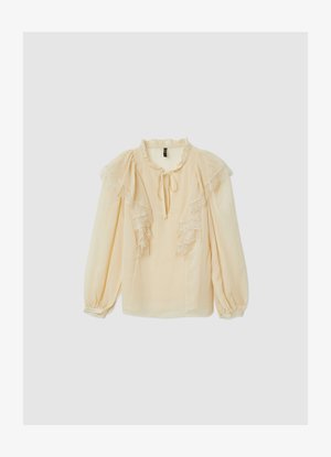 Blusa color beige chiaro con volant in pizzo e collo con laccetto. Presenta maniche lunghe con polsini arricciati e tessuto liscio, leggermente trasparente.