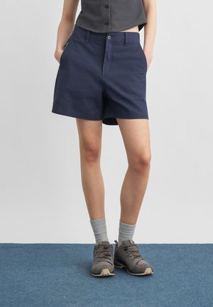 Person iført marineblå shorts, grå sokker og grå vandresko, stående på en blå tæppe mod en ensartet lys baggrund.