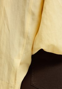 Κίτρινο υφαντό ύφασμα που κρέμεται πάνω από σκούρο καφέ ύφασμα με ορατή ραφή τσέπης και λεπτομέρειες ραφής.