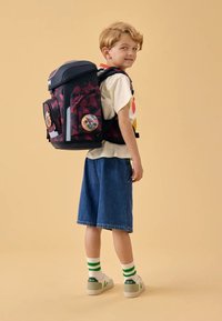 Mochila con un patrón geométrico azul oscuro y marrón, con dos parches circulares en la parte frontal. Niño viste pantalones cortos de mezclilla y calcetines a rayas con zapatillas.