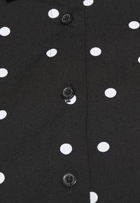 Czarna koszula z guzikami na całej długości, w białe kropki, gładki materiał, małe czarne guziki wzdłuż listwy guzikowej oraz starannie wykonany kołnierz.