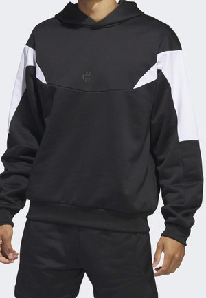 Man draagt een zwarte hoodie met witte strepen op de schouders en een klein logo op de borst, gecombineerd met een zwarte broek.