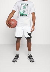 Bílý grafický tričko s zeleným potiskem, spárované s bílými šortkami s černým akcentem. Šedé sportovní tenisky a šedé ponožky. Drží basketbal.