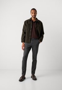 Hombre vestido con una camisa de overshirt abotonada de color verde oscuro, una camiseta tipo polo marrón, pantalones grises y mocasines marrones oscuros, de pie contra un fondo liso.