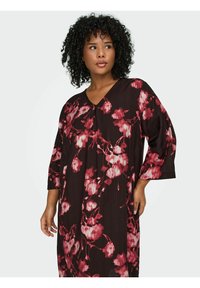 Cvjetna haljina od tamnosmeđeg materijala, s velikim uzorcima ružičastih i bijelih cvjetova. Dizajn s V izrezom i rukavima do tri četvrtine te detaljima nabora.