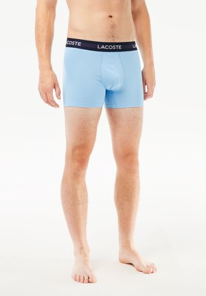 Boxer en stretch bleu clair avec une taille noire arborant le logo "LACOSTE". Texture lisse, design ajusté et longueur au-dessus du genou.