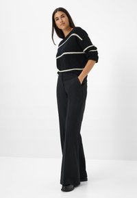 Un pull en tricot noir avec des rayures blanches horizontales, assorti à un pantalon large noir taille haute. Modèle portant des chaussures noires, les mains dans les poches.