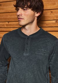Langarmhemd in Dunkelgrau, aus weichem Stoff gefertigt. Verfügt über einen knopfbaren Ausschnitt und ein kleines besticktes Logo auf der Brust.