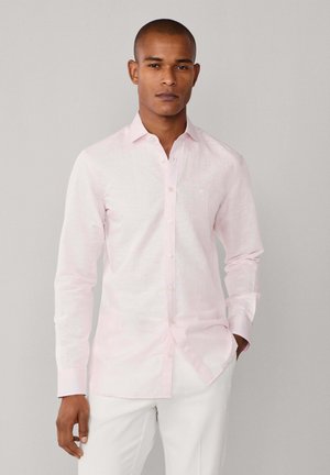 Homme portant une chemise à manches longues rayée rose clair et un pantalon blanc, debout avec une main dans la poche devant un fond gris uni.