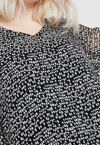 Personne blonde portant un chemisier noir transparent avec un motif de points irréguliers blancs et un décolleté en V, visible des épaules au milieu du torse.