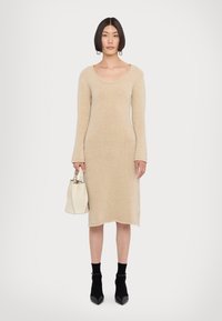 Abito lavorato beige con scollo a cerchio e maniche lunghe, abbinato a calzini neri alla caviglia e tacchi a punta neri; porta una piccola borsa bianca.