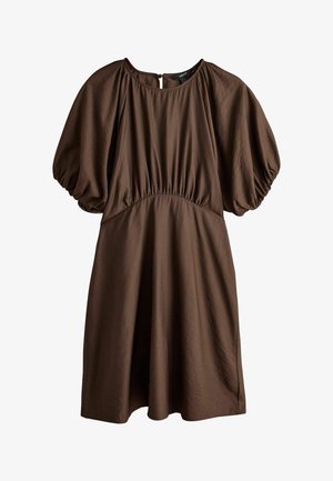 Jeune femme debout, mains sur les hanches, portant une robe marron ample avec des manches courtes bouffantes, devant un mur clair uni.
