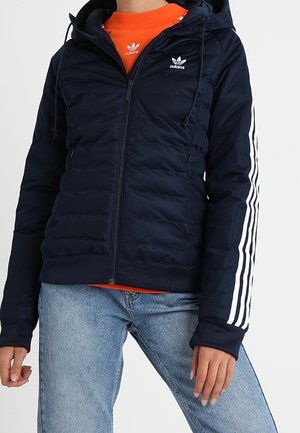 Veste matelassée Adidas de couleur bleue marine avec capuche, trois bandes blanches sur les manches, fermeture éclair à l'avant, portée par-dessus un sweat-shirt orange Adidas et un jean bleu.