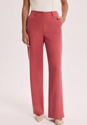 Kobieta nosząca różowe spodnie z wysokim stanem i prostymi nogawkami, z przodu zapięte na guziki i z bocznymi kieszeniami, w połączeniu z plecionymi butami na spiczastym nosku.