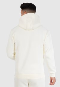 Sudadera con capucha color crema, con bolsillo tipo canguro, puños y dobladillo de canalé, hecha de una tela suave y texturizada. Presenta una capucha amplia.
