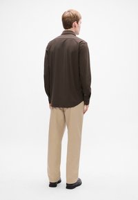 Camisa marrón de manga larga con collar, puños abotonados y un corte relajado, combinada con pantalones beiges y zapatos negros sobre un fondo liso.