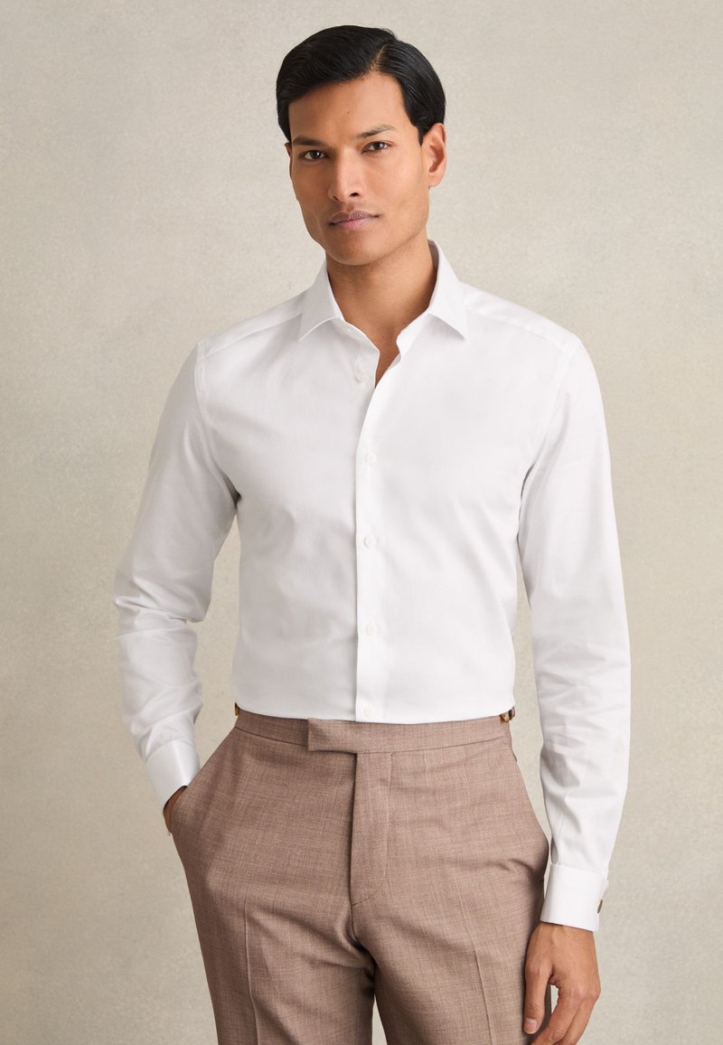 Homme portant une chemise blanche à manches longues boutonnée et un pantalon beige, debout, une main dans la poche, devant un fond uni.