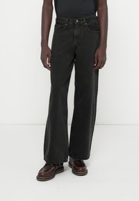 Μαύρες φαρδιές τζιν φόρμες από τζιν ύφασμα, με ψηλό μέσο, μπροστινές τσέπες και μίνιμαλ λεπτομέρειες ραφής. Συνδυασμένες με σκούρες παπούτσια.