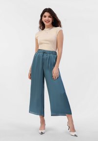 Beige ribbed short-sleeve top paired with wide-legged teal trousers and white heels. Trousers feature an elastic waistband and tie detail.