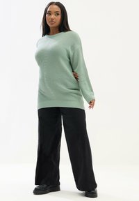 Maglione a maglia verde chiaro con una texture a costine, maniche lunghe e una vestibilità rilassata, abbinato a pantaloni neri a gamba larga e calzature nere chunky.